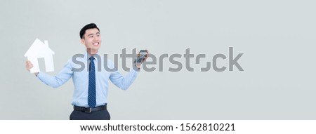 Young smiling Asian male real estate agent holding calculator and house cutout isolated on light gray banner background with copy space