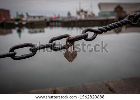 A romantic closed and rusty heart shaped padlock hanging from a chain beside a river with blurred cityscape in the background. Concept of long lasting and lost loves.