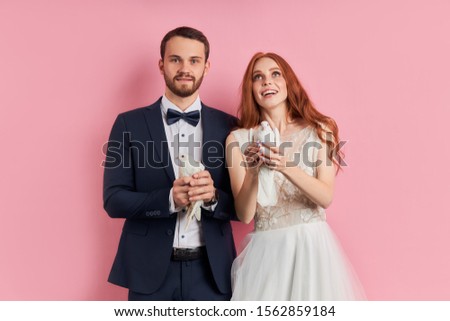 Attractive woman with red hair and man wearing tuxedo holding pigeons in hands, happy together, stand isolated over pink background . Dove symbol of happy family life