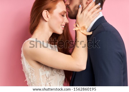 Closeup portrait of hugging and kissing couple, bearded man kiss forehead of woman with red hair isolated over pink background. Love concept