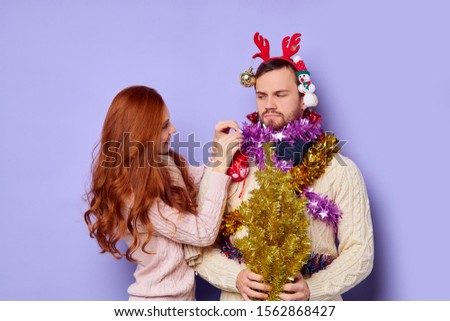 Dissatisfied, displeased man looking at chemung cheerful girl decorating him with different Christmas baubles, isolated family shut, posing over blue background, indoor shot
