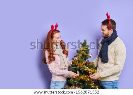 Happy girl and boy in red antler, looking at each others with lovely smile, waiting for guests with gifts, keeping hands on Christmas tree, isolated on violet background, indoor shot