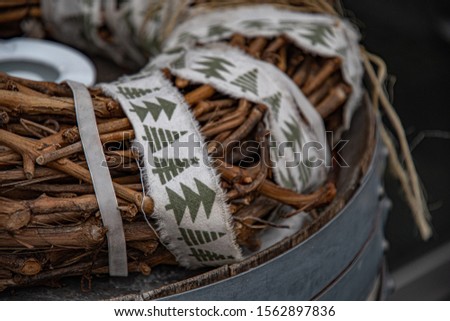 Christmas wreath made from woven brown grapevine branches and wrapped by twine and dirty white ribbons with primitive green pattern of Christmas trees. Top of old barrel with riveted metal hoops. 