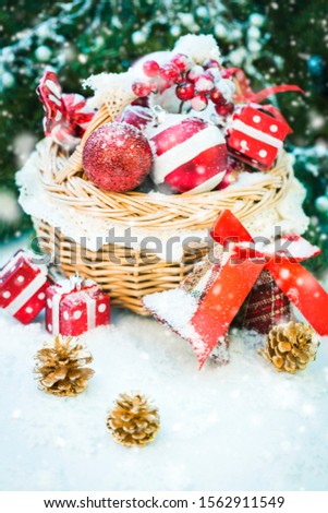 Richly filled Christmas basket with red festive balls, wrapped gifts, berries and pine cones on snowy background with snowing effect. Warm tones.