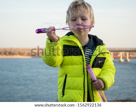 Funny blond child boy in bright yellow jacket walks in amusement park. Blonde kid blows soap bubbles from special ring on river background. Soapy foam in pink packaging. Good family vacation