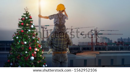 Asian boy on father's shoulders near Christmas tree with background of new high buildings and silhouette construction cranes of evening sunset, father and son on Christmas time