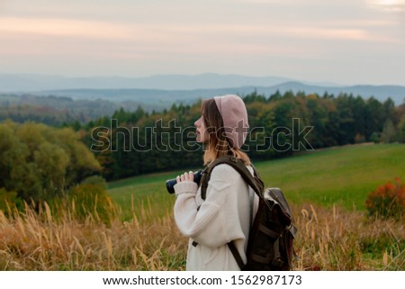Style girl with binoculars and backpack at countryside with mountains on background