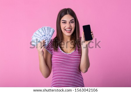 Excited attractive woman holding smartphone and money, cashback. Image of excited young lady isolated over pink background. Showing display of mobile phone holding money.