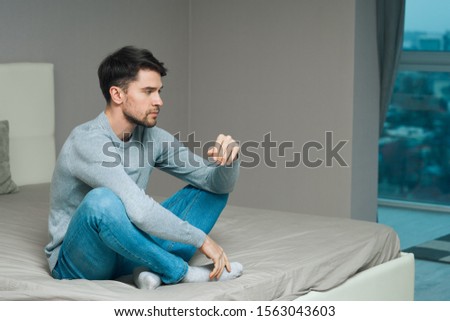 Side view of a man in jeans and a sweater sitting on a bed in a gray room