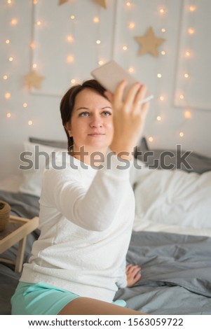 christmas, winter and people concept - happy young woman in bed taking selfie on smartphone at home bedroom