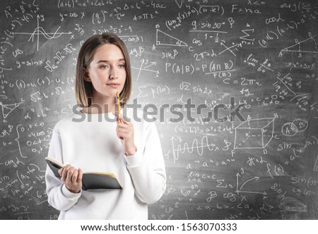 Thoughtful young woman in casual clothes with notebook standing near blackboard with math formulae written on it. Concept of education and science Royalty-Free Stock Photo #1563070333