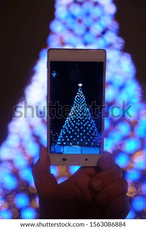 man holds in his hand new model of  smartphone and take pictures of garlands and Christmas lights on New Year tree. Christmas Eve. take photo. 