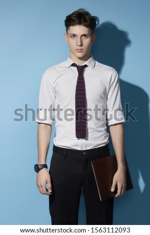 Medium full shot of a young dark haired man in a black trousers and white printed shirt with a deep blue knit tie with burgundy stripes. The man with a black watch is holding a leather folder. 