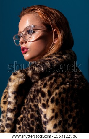 Attractive girl with blue eyes, red hair, trendy glasses and in fur leopard coat. Studio portrait.