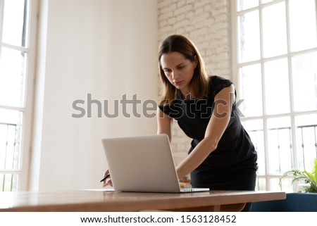 Worried young businesswoman leaning over table, typing answer in computer to email in hurry at workplace. Puzzled female company employee worker manager looking at laptop monitor, writing notes.