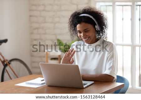 Happy african american young business woman wearing wireless headphones, looking at computer screen, waving hello. Pleasant attractive mixed race female professional holding video call with client. Royalty-Free Stock Photo #1563128464