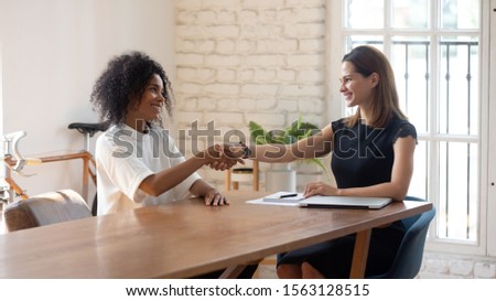 Happy african american young female job applicant shaking hands with pleasant hr manager at office, celebrating employment. Two multiracial business women making agreement at meeting at workplace. Royalty-Free Stock Photo #1563128515
