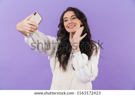 Image of cheerful pleased cute young brunette woman posing isolated over purple wall background take a selfie by mobile phone waving talking.
