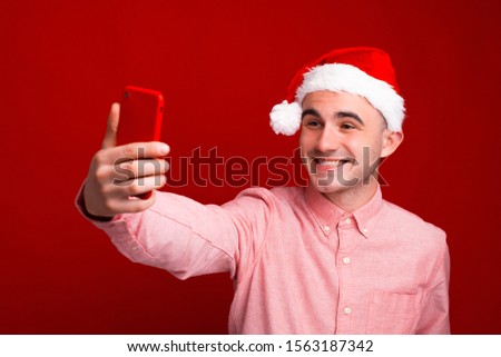 Photo of happy young man wearing santa claus hat and taking selfie with smartphone