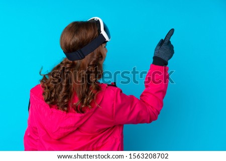 Skier woman with snowboarding glasses over isolated blue wall pointing back with the index finger