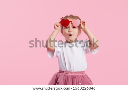 Portrait of surprised cute little toddler girl in the heart shape sunglasses. Child with open mouth having fun isolated over pink background. Looking at camera. Wow funny face Royalty-Free Stock Photo #1563226846