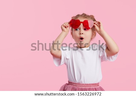 Portrait of surprised cute little toddler girl in the heart shape sunglasses. Child with open mouth having fun isolated over pink background. Looking at camera. Wow funny face Royalty-Free Stock Photo #1563226972