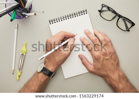 cropped view of man writing in notebook near glasses and stationery isolated on grey 