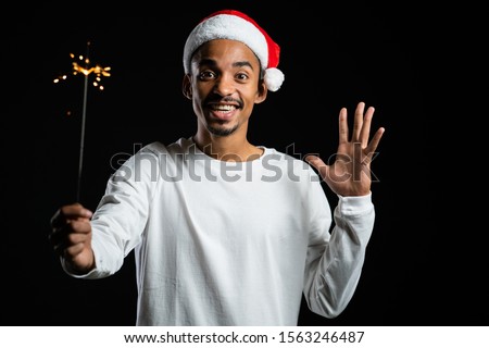 African american man in Santa hat celebrating with sparkler and bengal fire. Happy mix race guy smiling and having fun against black background
