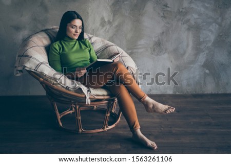 Photo of interested woman reading book in armchair pants orange being barefoot isolated grey wall color concrete background in spectacles