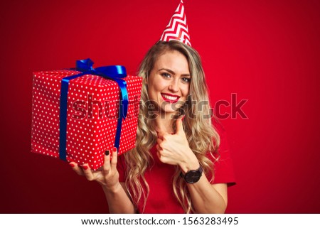 Young beautiful woman holding bitrhday gift over red isolated background happy with big smile doing ok sign, thumb up with fingers, excellent sign