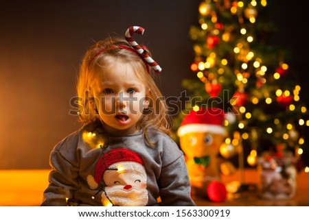 Unhappy caucasian blonde baby girl, sitting in front of Christmas tree, displeased facial expression, Happy New Year 2022 celebration holiday concept 