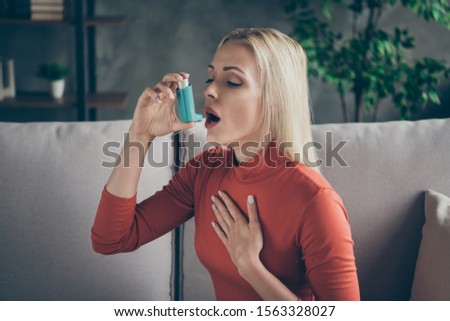 Profile photo of unhealthy pretty blond lady suffering allergy holding inhaler breathing in feel better sitting couch wear orange pullover shirt living room indoors