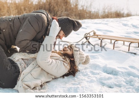 Happy winter couple smiling having fun, fooling around like children in winter park near a snowy river. Young beautiful woman and handsome man in winter warm casual clothes laying on snow playing in
