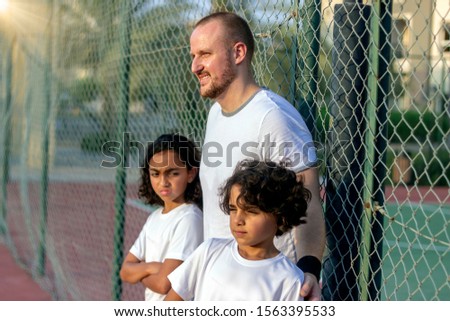 A young coach and kids are resting during basketball game.The kids are standing in the schoolyard.Two adorable young kids with their coach standing against a fence on basketball court and looking away