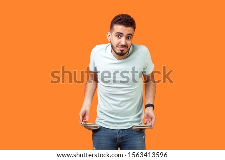 Portrait of frustrated worried brunette man with beard in casual white t-shirt turning out empty pockets showing I have no money gesture, bankrupt. indoor studio shot isolated on orange background Royalty-Free Stock Photo #1563413596