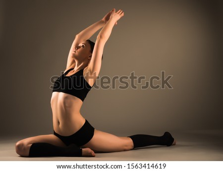Beautiful young healthy girl in sportswear does a workout for the joints of the legs while sitting on the studio floor in dark lighting. Healthy lifestyle concept. Advertising space