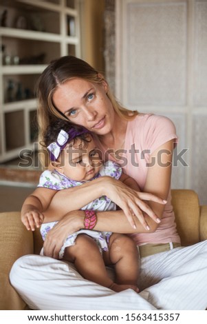 Attractive young white mother hugging her toddler daughter. A woman is sitting on a soft chair indoors. Multiracial family.