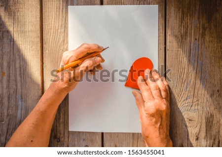 Elderly woman hand going to write a letter, red heart on rustic wooden table as a background, sun rays. View from above, copy space. Mother or Woman, Valentine day, human emotions,  warmth.