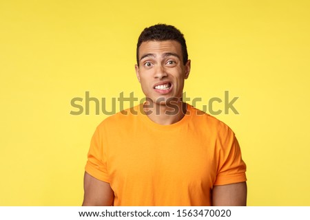 Yikes, awkward. Embarrassed young handsome man caught on lie, feel indesicive and slightly worried, cringe, smiling nervously and look camera with reluctance, stand yellow background Royalty-Free Stock Photo #1563470020