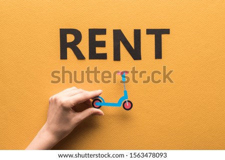 cropped view of woman holding paper cut kick scooter near rent lettering on orange background