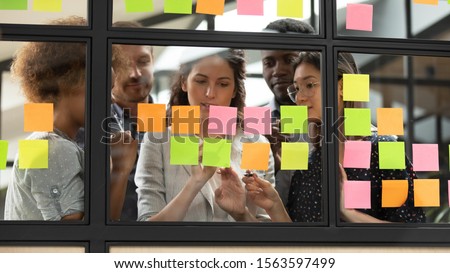 Focused young diverse teammates working with caucasian team leader on IT startup project kanban organization process, discussing details and tasks, writing notes on colorful paper stickers at office. Royalty-Free Stock Photo #1563597499