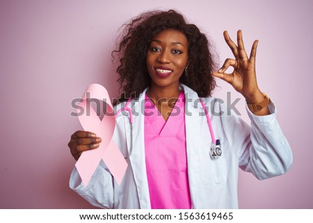 Young african american doctor woman holding cancer ribbon over isolated pink background doing ok sign with fingers, excellent symbol