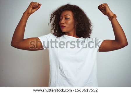 Young african american woman wearing t-shirt standing over isolated white background showing arms muscles smiling proud. Fitness concept.