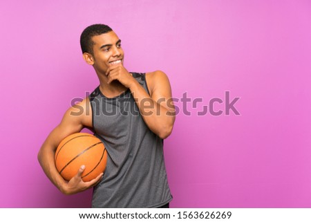 Young sport man with ball of basketball over isolated purple wall thinking an idea and looking side