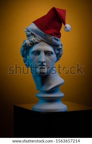 White gypsum Statue of Apollo Belvedere in a red cap of Santa Claus in blue contour light on colorful backgrounds.