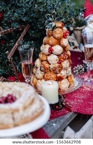 Winter, christmas decor: glasses of champaign, candles, delicious french desserts - profiterole and paris-brest, santa clause. Red festive table cloth, white snow. Green holly bush on background