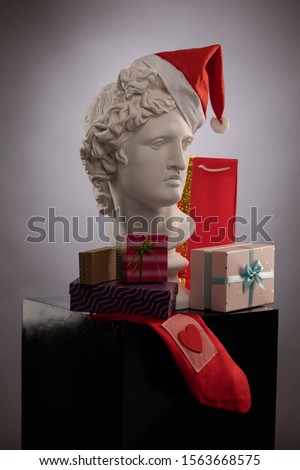White plaster Statue of Apollo Belvedere in a red cap of Santa Claus, gifts boxes and packages on colorful backgrounds. Composition for congratulations, background for design.