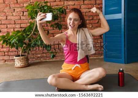 Photo of smiling young woman with red hair taking selfie photo on cellphone while sitting on mat in room