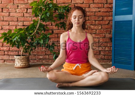 Image of a beautiful young fitness redhead woman meditate indoors over brick wall and tree plant background.
