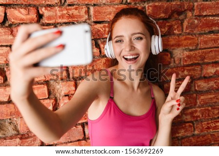 Photo of pleased woman in headphones taking selfie photo on cellphone while gesturing peace sign isolated over brick wall indoors